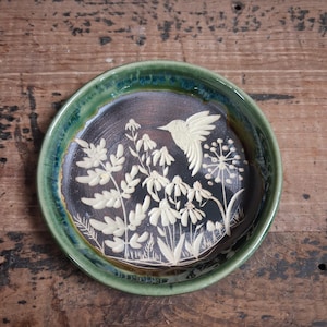 May include: A round ceramic dish featuring a dark brown center with a cream-colored relief design of a hummingbird hovering over wildflowers. The dish has a raised, glossy green rim with subtle blue-toned glaze variations along the edge.