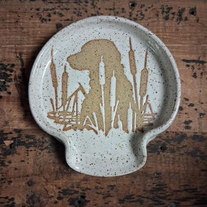 May include: A speckled, off-white ceramic spoon rest with a brown dog silhouette and cattail design. The spoon rest has a curved shape with a small lip for holding utensils. The design is rustic and nature-themed.