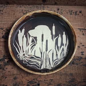 May include: A round, ceramic plate with a dark brown interior and a lighter brown rim. The plate features a cream-colored silhouette of a dog in a marsh setting with cattails and water.