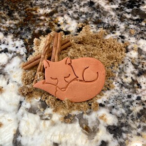 May include: A terracotta-colored fox cookie cutter, shaped like a sleeping fox, is nestled in brown sugar.  Cinnamon sticks are visible in the background.