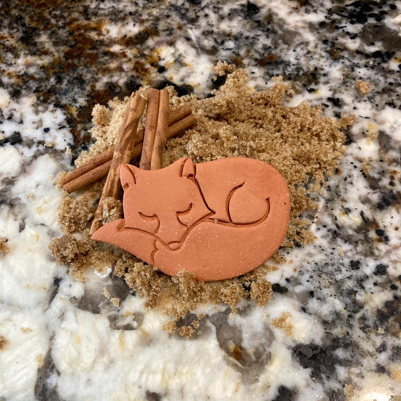 May include: A terracotta-colored fox cookie cutter, shaped like a sleeping fox, is nestled in brown sugar.  Cinnamon sticks are visible in the background.