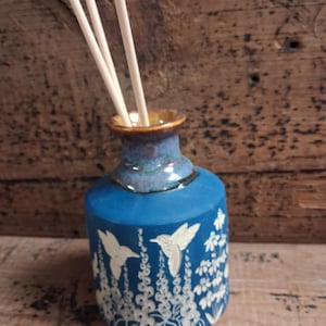 May include: A blue ceramic essential oil diffuser with a hummingbird and floral design. The diffuser has a narrow neck and a wide base, with three wooden reeds inserted. The background is a rustic wooden surface.