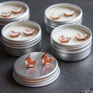 May include: Five small silver tins with lids, each containing a pair of wooden fox earrings. The foxes are painted in shades of white, orange, and pink.