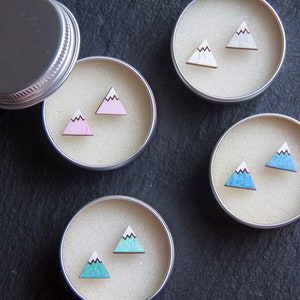 May include: Four small round containers with lids, each containing a pair of wooden earrings shaped like mountains. The earrings are painted in different colours: pink, white, blue, and turquoise.