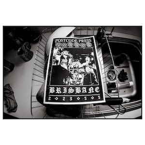 May include: Black and white poster for "Postcode Press" with a band performing. The word "Brisbane" is printed in large gothic letters. The poster is resting in a metal dish rack next to a kitchen sink.