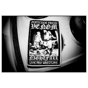 May include: Black and white photo of a sticker advertising a live pro wrestling event called "Nightfall" featuring the band Venom. The sticker is on a white surface and has the text "Postcode Press Venom Nightfall Live Pro Wrestling" on it.