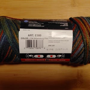 May include: A skein of Red Heart yarn in the Williamsburg Print colorway. The yarn is a blend of blue, green, and orange hues. The label displays the product details, including the art number E300 and color code 0788.