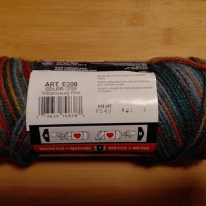 May include: A skein of Williamsburg Print yarn, featuring a blend of colors including teal, orange, and gold. The label displays the product details: ART. E300, COLOR: 0788, and WORSTED - MEDIUM. Ideal for knitting and crochet projects.