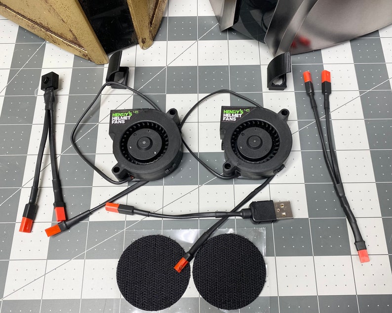 Puede incluir: Dos ventiladores de casco negros con texto verde y blanco que dice "Henry's Helmet Fans". Los ventiladores est&aacute;n conectados a cables negros con conectores rojos. Se incluyen dos almohadillas adhesivas negras.