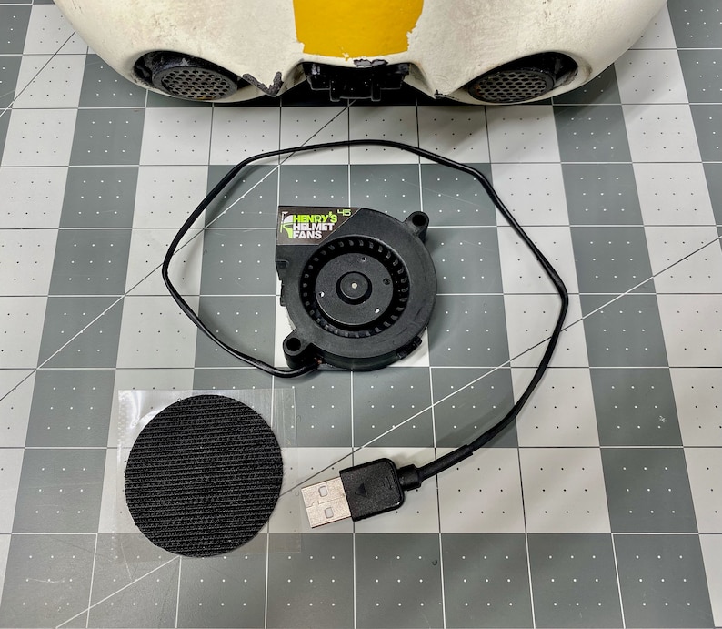 May include: A black helmet fan with a USB cable, a black velcro pad, and a white helmet with a yellow stripe. The fan has the text "HENRY'S HELMET FANS" printed on it.