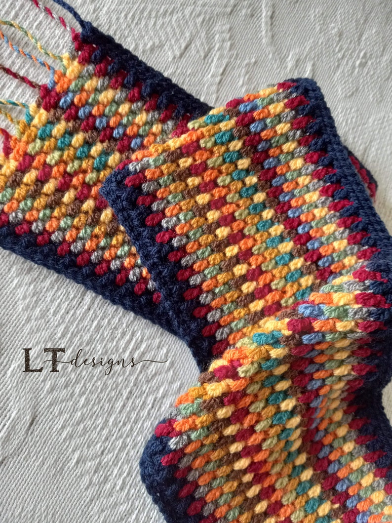 May include: A colorful crocheted scarf with a navy blue border and fringe. The scarf is made with a variety of colors, including red, orange, yellow, green, blue, and brown. The scarf is folded in half and the fringe is visible at the top of the image. The text "LT Designs" is visible in the lower left corner of the image.