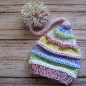 May include: A striped knitted hat with a long pink pom-pom. The hat is made in pastel colors of pink, yellow, green, blue, and white.