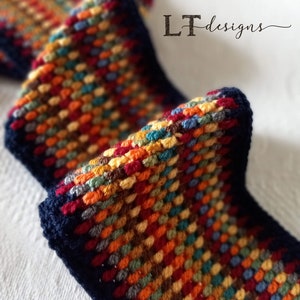 May include: A close-up of a colorful crocheted scarf with a navy blue border. The scarf is made with a variety of colors, including red, orange, yellow, green, blue, and brown. The scarf is folded in half and the ends are tucked in.