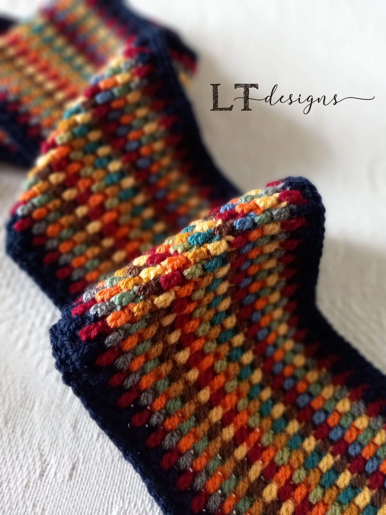 May include: A close-up of a colorful crocheted scarf with a navy blue border. The scarf is made with a variety of colors, including red, orange, yellow, green, blue, and brown. The scarf is folded in half and the ends are tucked in.