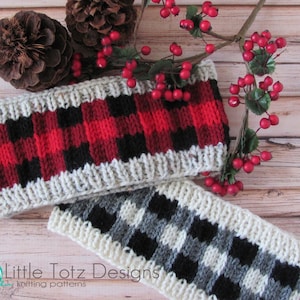 May include: Two knitted headbands with a plaid pattern. The top headband is red, black, and white. The bottom headband is black, gray, and white. The headbands are on a wooden background with a sprig of red berries and pine cones.