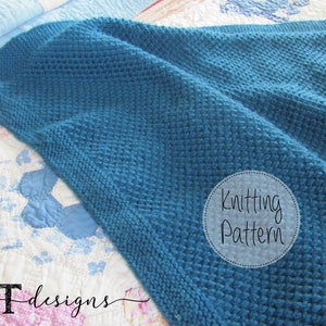 May include: A teal blue knitted blanket with a textured stitch pattern. The blanket is folded on a white and blue quilt. The text "Knitting Pattern" is written in a circle on the blanket.