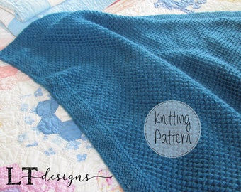 May include: A teal blue knitted blanket with a textured stitch pattern. The blanket is folded on a white and blue quilt. The text "Knitting Pattern" is written in a circle on the blanket.