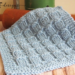 May include: A light blue knitted dishcloth with a textured pattern. The dishcloth is lying on a wooden surface.