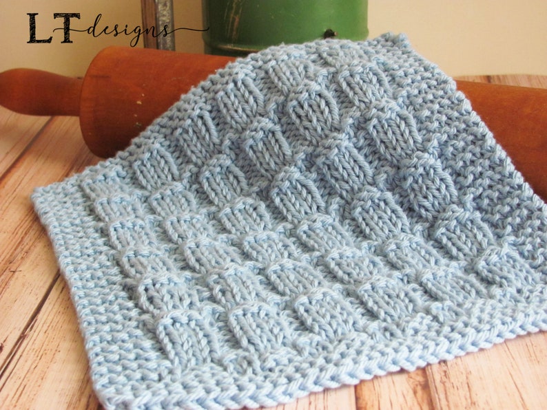 May include: A light blue knitted dishcloth with a textured pattern. The dishcloth is lying on a wooden surface.