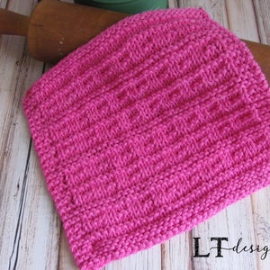 May include: A pink knitted dishcloth with a textured pattern. The dishcloth is lying on a wooden surface.