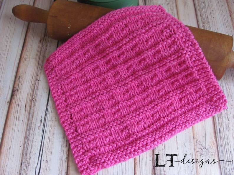 May include: A pink knitted dishcloth with a textured pattern. The dishcloth is lying on a wooden surface.