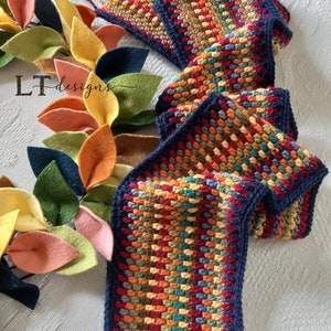 May include: A colorful crocheted scarf with a navy blue border and fringe. The scarf is made with a variety of colors, including red, orange, yellow, green, and blue. The scarf is lying on a white surface with a garland of felt leaves.