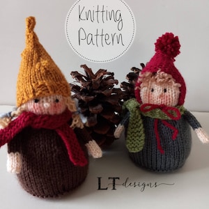 Könnte beinhalten: Strickmuster für zwei niedliche Gnome, einer mit einem braunen Hut und einem roten Schal, der andere mit einem roten Hut und einem grünen Schal. Die Gnome sitzen auf einer weißen Oberfläche mit einer Tannzapfe im Hintergrund.