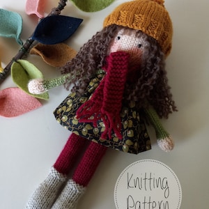 May include: Knitting pattern for a doll with brown curly hair, wearing a red scarf, a brown dress with a floral pattern, a mustard yellow hat, and grey knitted boots. The doll is surrounded by felt leaves in various colors.