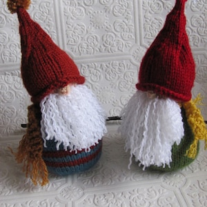 May include: Two knitted gnomes with red hats, white beards, and colourful striped bodies. The gnome on the left has a brown yarn beard and the gnome on the right has a green body with a yellow scarf.