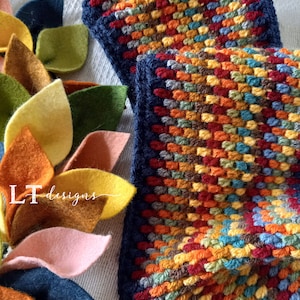 May include: A close-up of a colorful crocheted scarf with a blue border. The scarf is made with a variety of colors, including red, orange, yellow, green, and blue. The scarf is lying on a white surface with felt leaves.