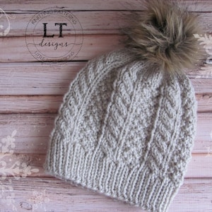May include: A beige knitted hat with a cable knit pattern and a fluffy brown pom pom. The hat is on a wooden background with a white snowflake design.