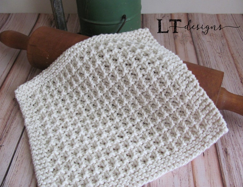 May include: A white knitted dishcloth with a textured pattern. The dishcloth is lying on a wooden surface.
