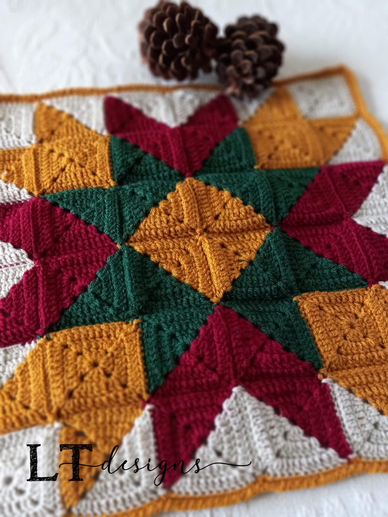 May include: A crocheted square with a star design in shades of red, green, gold, and white. The square is framed with a brown border. The text "LT Designs" is visible in the bottom left corner.