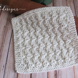 May include: A white knitted dishcloth with a textured diamond pattern. The dishcloth is lying on a wooden surface.