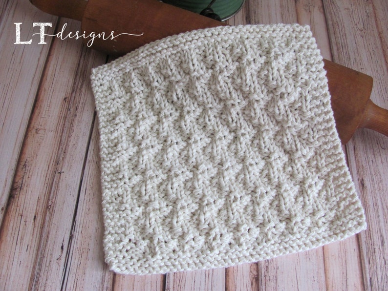 May include: A white knitted dishcloth with a textured diamond pattern. The dishcloth is lying on a wooden surface.