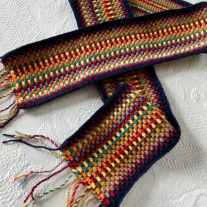 May include: A colorful crocheted scarf with a navy blue border and fringe. The scarf is made with a variety of colors, including red, orange, yellow, green, blue, and brown. The scarf is folded in half and the fringe is hanging down on both sides. The scarf is on a white surface. LT designs.