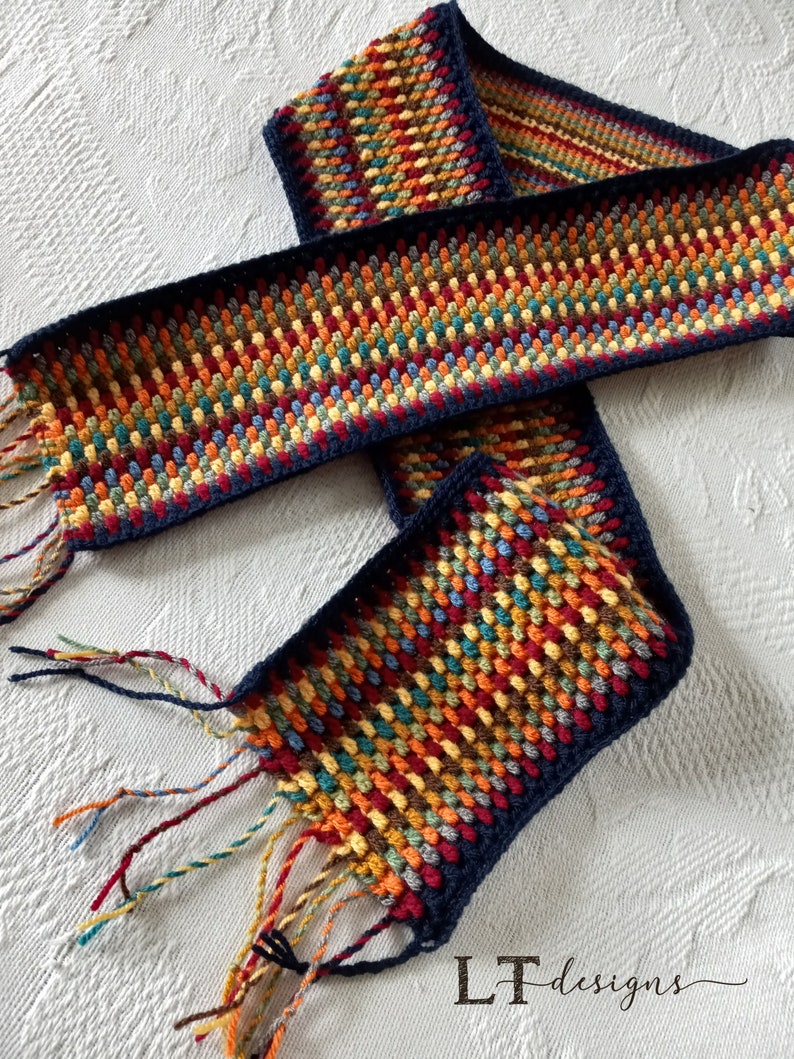 May include: A colorful crocheted scarf with a navy blue border and fringe. The scarf is made with a variety of colors, including red, orange, yellow, green, blue, and brown. The scarf is folded in half and the fringe is hanging down on both sides. The scarf is on a white surface. LT designs.