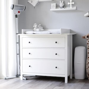 Peut inclure: Une chambre d'enfant avec une commode blanche à trois tiroirs et boutons noirs, un grand ours en peluche blanc, une pile de cubes de rangement transparents, un tapis rond blanc, une girafe en peluche et deux étagères murales blanches décorées.