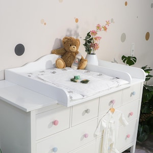 Pode incluir: Uma cômoda branca com trocador, ursinho de pelúcia e mamadeira. A cômoda tem várias gavetas com puxadores coloridos. Um macacão branco pendurado na lateral. Um vaso de flores rosas está sobre a mesa.