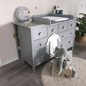 Op de afbeelding: Een grijze houten commode met acht lades voorzien van wolkvormige knoppen. Op het bovenblad liggen een aankleedkussen, een maanvormig kussen en een flesje. Een wit kledingstuk met lange mouwen hangt aan een ladeknop en een pluche olifant staat ernaast.