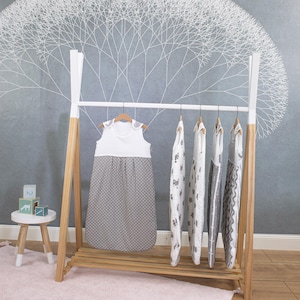 May include: A white wooden clothes rack with five grey and white baby sleeping bags hanging on it. The sleeping bags have different patterns, including polka dots, stars, and geometric designs. The rack is standing on a light pink rug.