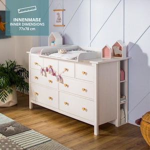 May include: A white wooden changing table dresser with seven drawers featuring gold heart-shaped knobs. The top surface includes a changing tray with a pad. Text on the image indicates inner dimensions of 77x78 cm. Side shelving is attached.