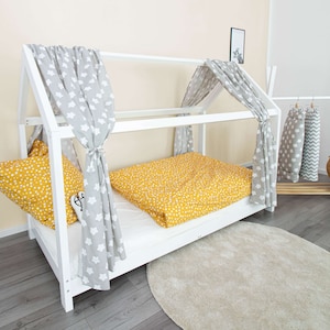 Könnte beinhalten: Ein weißes Hausbett aus Holz mit Matratze, gelber Bettwäsche mit weißen Punkten und grauen Vorhängen mit Sternenmuster. Das Bett hat einen Baldachinrahmen und steht auf einem hellgrauen Boden. Vor dem Bett liegt ein runder, beiger Teppich.