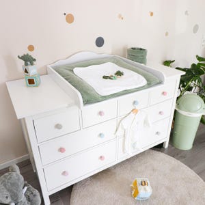 Op de afbeelding: Een witte houten ladekast met meerdere lades voorzien van pastelkleurige ronde knoppen. Bovenop ligt een saliegroen aankleedkussen met een wit doekje, naast een klein groen stoffen mandje en een stapel van twee houten blokken met een groene dinosaurus.