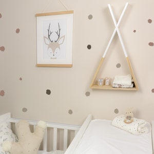 Op de afbeelding: Een babykamer met een wit wiegje, een witte commode en een houten tipi-rek. De muren zijn versierd met bruine en roze stippen-muurstickers. Een ingelijst printje van een hert met gewei hangt aan de muur.