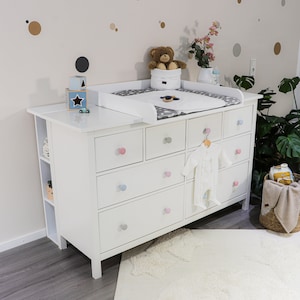 Puede incluir: Una cómoda blanca para bebés con múltiples cajones y pomos coloridos. Una colchoneta para cambiar pañales con un patrón gris y blanco se encuentra encima, junto con un oso de peluche y ropa de bebé. La habitación está decorada con calcomanías de pared y una alfombra mullida.
