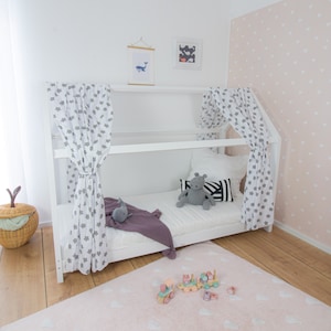 Könnte beinhalten: Weißes, hausförmiges Bettgestell aus Holz mit einer weißen Matratze und grauen und weißen Sternenmustervorhängen. Das Bett ist mit einer lilafarbenen Decke, einem grauen Stofftier und einem schwarz-weiß gestreiften Kissen dekoriert. Das Bett steht auf einem hellen Holzboden mit einem rosa Teppich mit weißen Herzen.