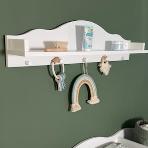 Op de afbeelding: Witte wandplank met een gebogen bovenkant, voorzien van een babylotionfles, een houten haarborstel, een klein potje crème en drie haken. Een regenboogkleurige macramé-hanger en een houten speelgoed hangen aan de haken.