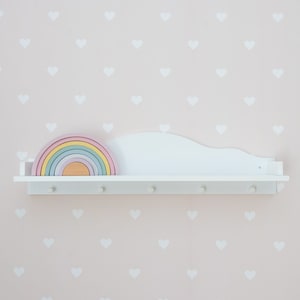 Peut inclure: Étagère en bois blanc avec un dos en forme de nuage et des anneaux d'empilage en bois aux couleurs de l'arc-en-ciel. L'étagère a cinq crochets et est montée sur un mur rose avec des autocollants en forme de cœur blancs.