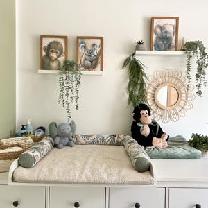 Op de afbeelding: Een witte commode met een beige textuurkussen, een kussen met palmbladpatroon, een grijze olifantenknuffel en een zwarte apenknuffel. Boven de commode hangen twee witte planken met ingelijste dierenprints en een ronde rotan spiegel.
